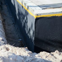 MICHAŁ GALAS GALASBRUK - Zabezpieczona ściana fundamentowa w wykopie, pokryta czarną izolacją z żółtą taśmą uszczelniającą, widoczne ślady cienia drzewa.