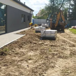 Dwa żółte mini-koparki na placu budowy przed nowoczesnym domem z dużymi oknami, palety z kostką brukową i robotnicy w oddali.