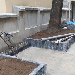 MICHAŁ GALAS GALASBRUK - Kaskadowe rabaty z szarej kostki obłożone ziemią, obok drzewo i betonowy murek z wnękami, na ziemi leży rura PCV i drewniane deski.