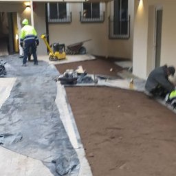 MICHAŁ GALAS GALASBRUK - Przygotowanie podłoża pod nawierzchnię w wewnętrznym patio budynku wielorodzinnego, widoczni pracownicy, ubijarka wibracyjna i rozplantowana warstwa gruntu.