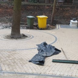 MICHAŁ GALAS GALASBRUK - Układanie beżowo-szarej kostki brukowej w okrągły wzór wokół drzewa, z widocznymi czarnymi workami, szczotką i elementami kanalizacji liniowej, częściowo ukończone prace.