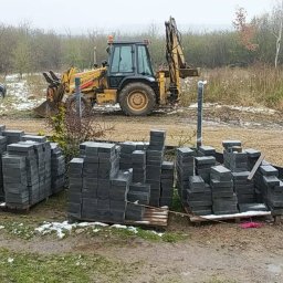 MICHAŁ GALAS GALASBRUK - Stosy szarych kostek brukowych na paletach w błotnistym terenie z żółtą koparką w tle, częściowo ośnieżone podłoże, widoczni pracownicy w odblaskowych kamizelkach i zaparkowany SUV.