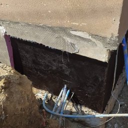MICHAŁ GALAS GALASBRUK - Zabezpieczony fundament budynku z widoczną hydroizolacją, wykopem i wystającymi rurami instalacyjnymi