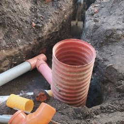 MICHAŁ GALAS GALASBRUK - Pracownik w zielonej czapce kopie dół pod rury kanalizacyjne za pomocą szpadla. W wykopie widoczna czerwona rura karbowana, obok różowe i szare rury oraz żółte odcinki rur karbowanych.