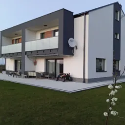 Nowoczesny dwupiętrowy dom z szaro-białą elewacją i tarasem wyłożonym jasną kostką brukową, z meblami ogrodowymi i dziecięcym pojazdem elektrycznym, widziany z perspektywy trawnika z młodym...