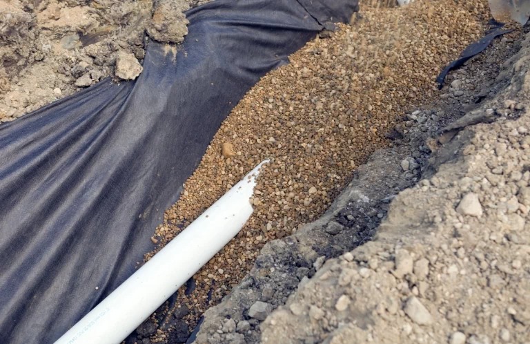 Wykop wyłożony czarną geowłókniną, wypełniony drobnym żwirem z ułożoną białą rurą drenarską z widocznym uszkodzeniem na końcu.