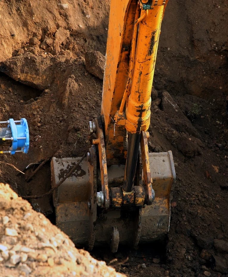 Żółta koparka zagłębia się łyżką w ziemię, obok widoczny fragment niebieskiej rury.