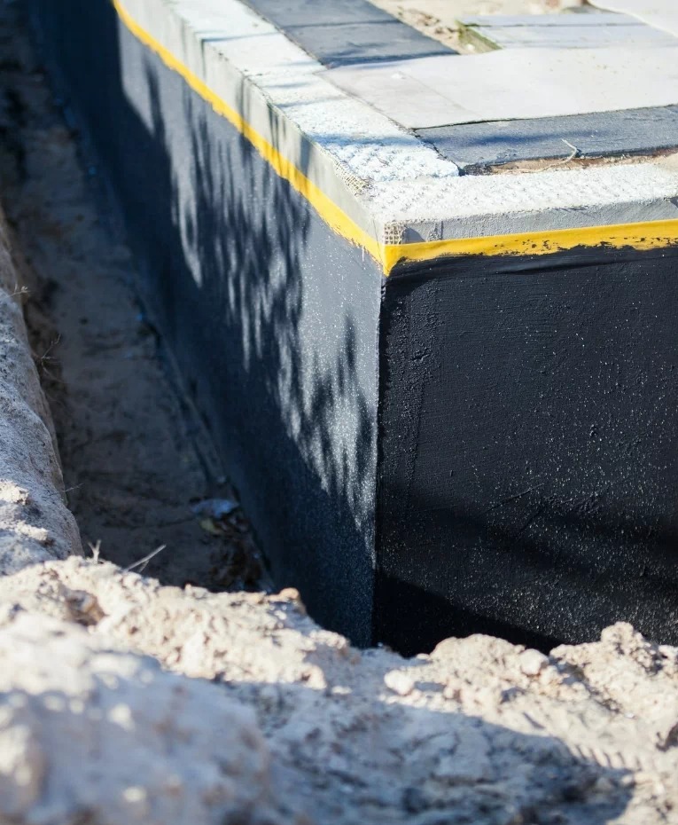 Zabezpieczona ściana fundamentowa w wykopie, pokryta czarną izolacją z żółtą taśmą uszczelniającą, widoczne ślady cienia drzewa.