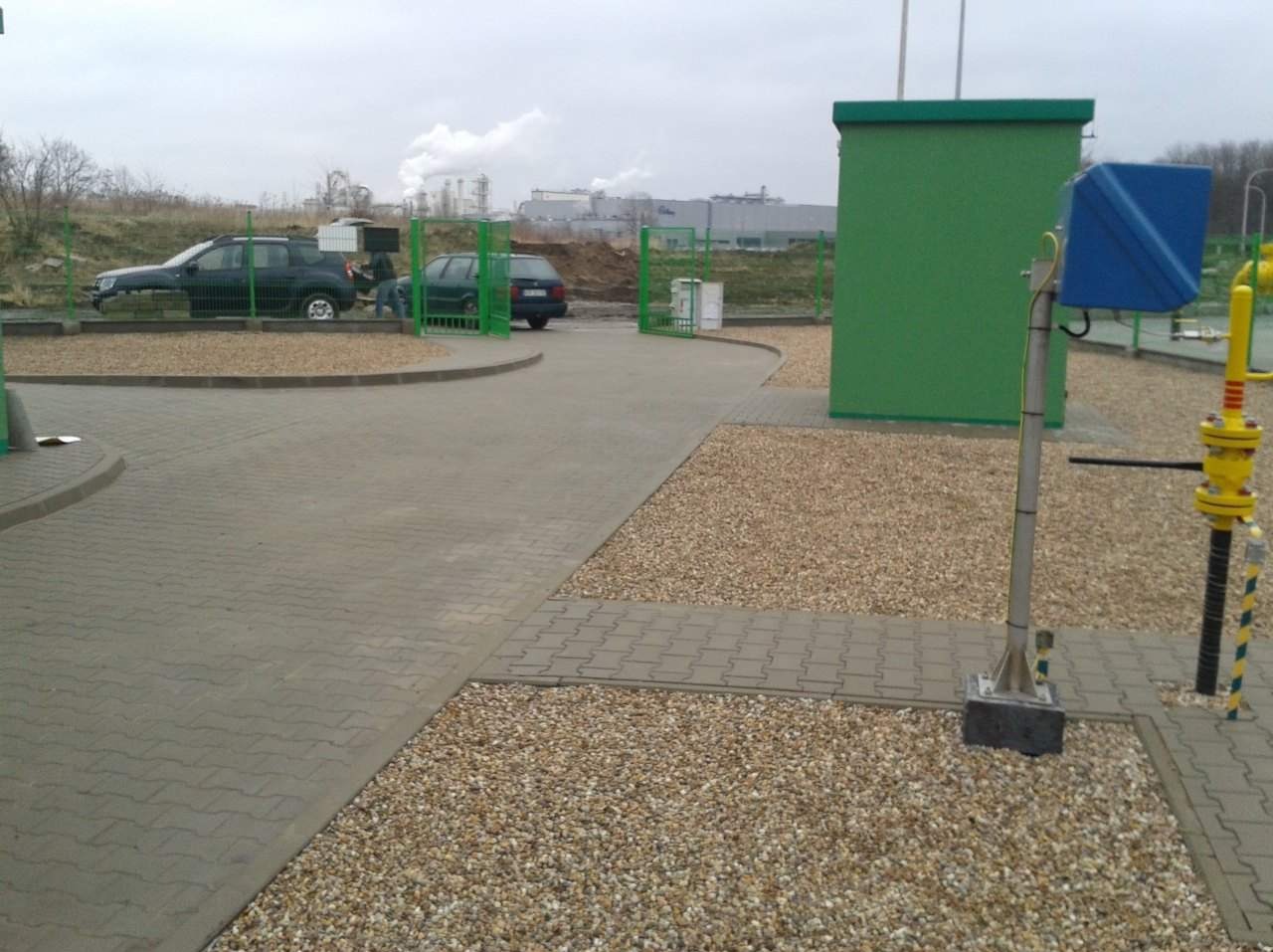 Ułożona szara kostka brukowa tworząca plac manewrowy, obok wysypany żwir, w tle zabudowania przemysłowe z kominami.