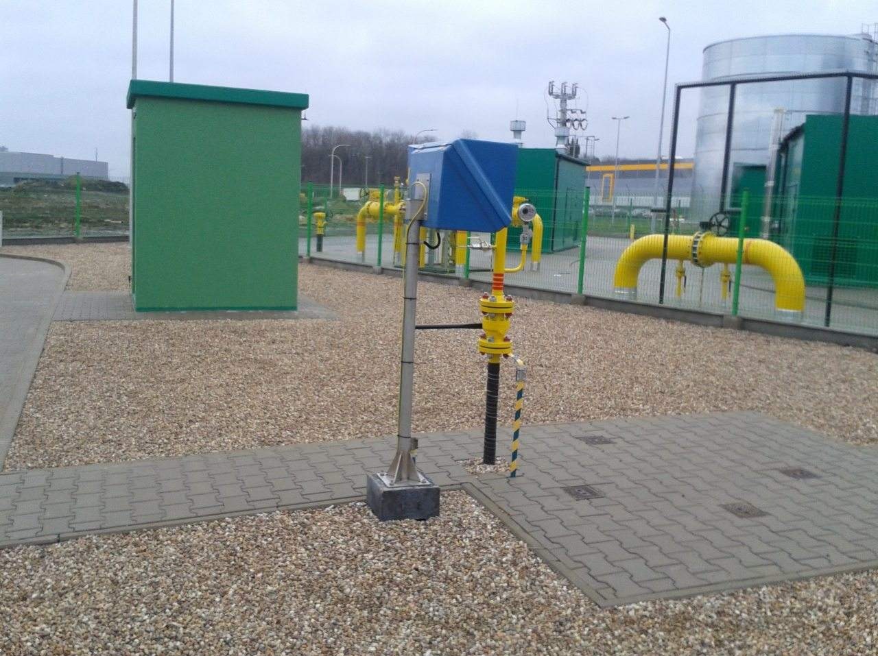 Instalacja gazowa z żółtymi rurami i niebieską obudową w Krakowie, otoczona żwirem i kostką brukową, z widocznymi elementami infrastruktury przemysłowej w tle.