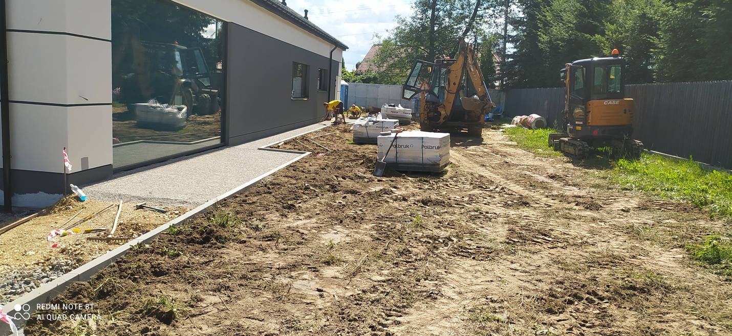 Dwa żółte mini-koparki na placu budowy przed nowoczesnym domem z dużymi oknami, palety z kostką brukową i robotnicy w oddali.