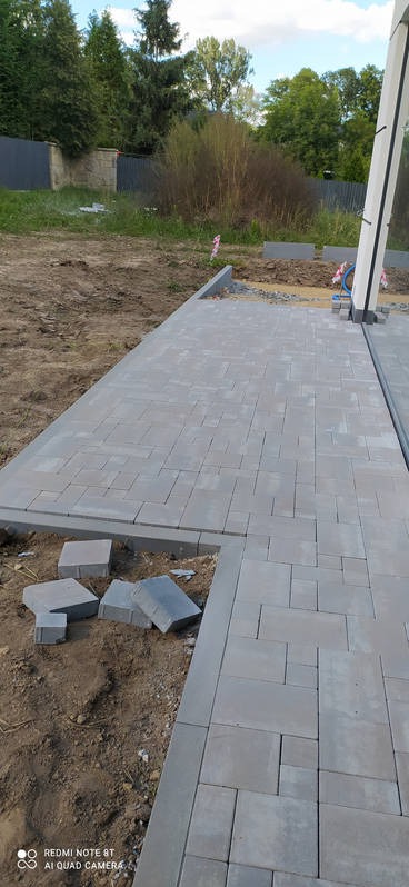 Fragment nowej szarej nawierzchni z betonowej kostki, z widocznymi niedokończonymi krawędziami i stertą kostek obok, na tle trawnika i drzew.