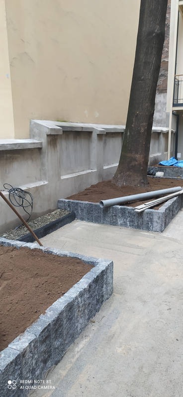 Kaskadowe rabaty z szarej kostki obłożone ziemią, obok drzewo i betonowy murek z wnękami, na ziemi leży rura PCV i drewniane deski.