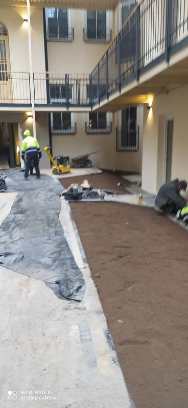 Przygotowanie podłoża pod nawierzchnię w wewnętrznym patio budynku wielorodzinnego, widoczni pracownicy, ubijarka wibracyjna i rozplantowana warstwa gruntu.