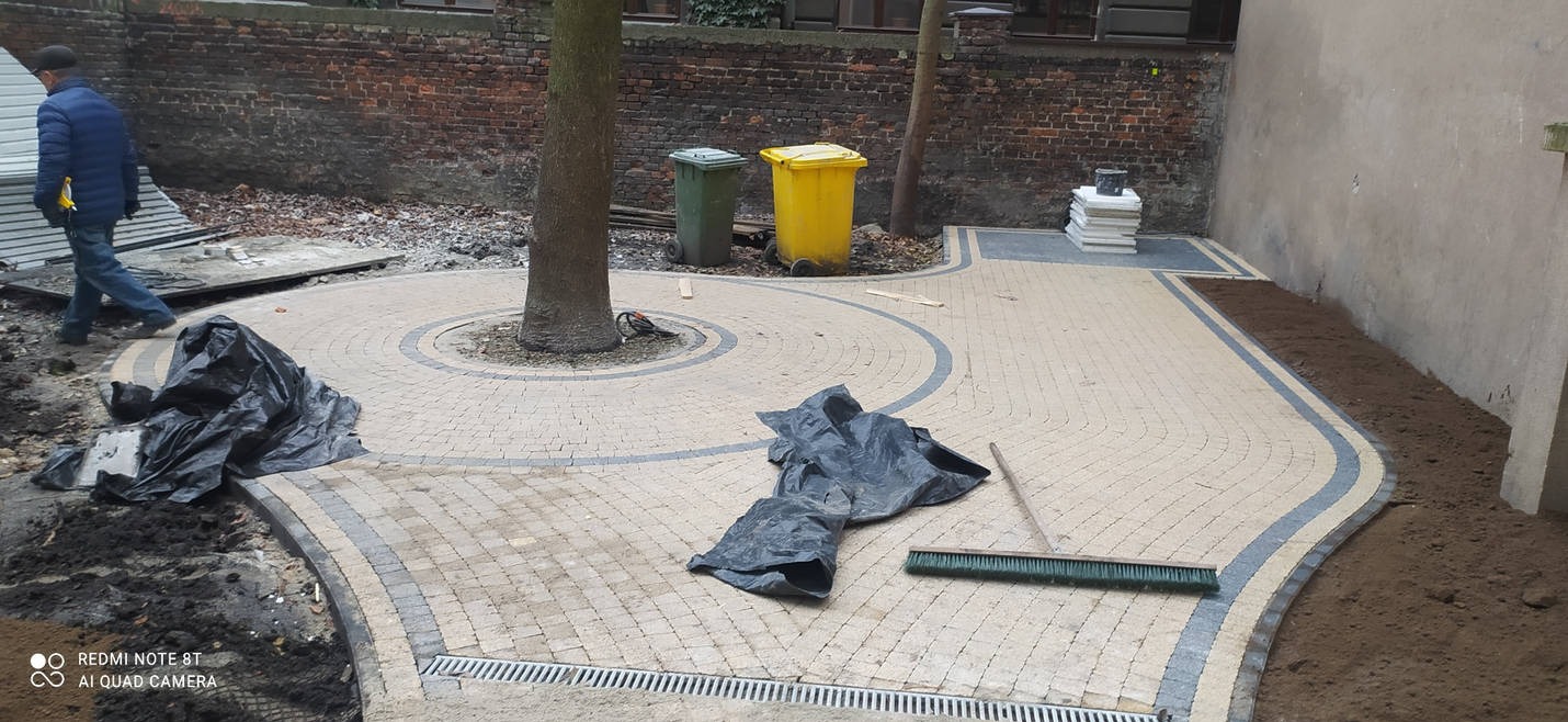 Układanie beżowo-szarej kostki brukowej w okrągły wzór wokół drzewa, z widocznymi czarnymi workami, szczotką i elementami kanalizacji liniowej, częściowo ukończone prace.