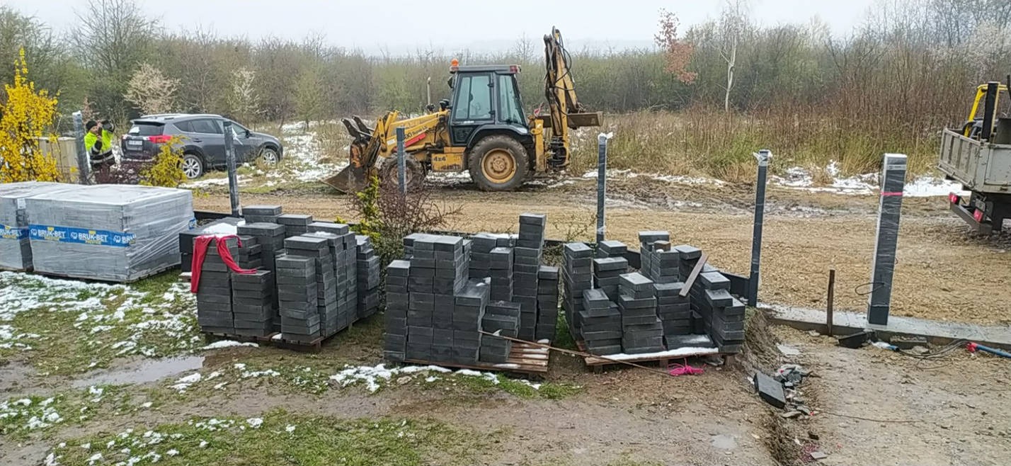 Stosy szarych kostek brukowych na paletach w błotnistym terenie z żółtą koparką w tle, częściowo ośnieżone podłoże, widoczni pracownicy w odblaskowych kamizelkach i zaparkowany SUV.