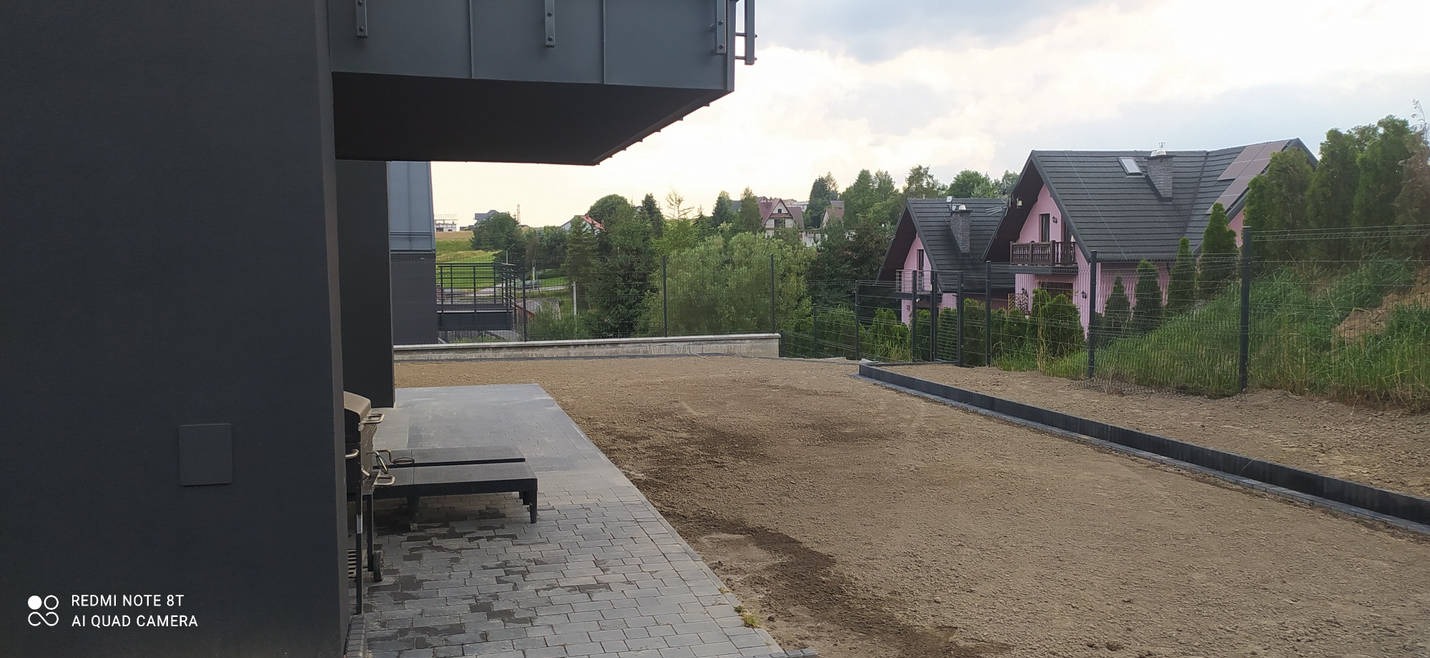 Przygotowany teren pod przyszłą nawierzchnię, widoczne obrzeża, częściowo ułożona kostka brukowa, w tle domy jednorodzinne.