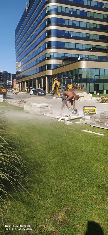 Pracownik tnie szlifierką kątową betonową kostkę na placu budowy przed nowoczesnym budynkiem biurowym, w tle widoczna koparka.