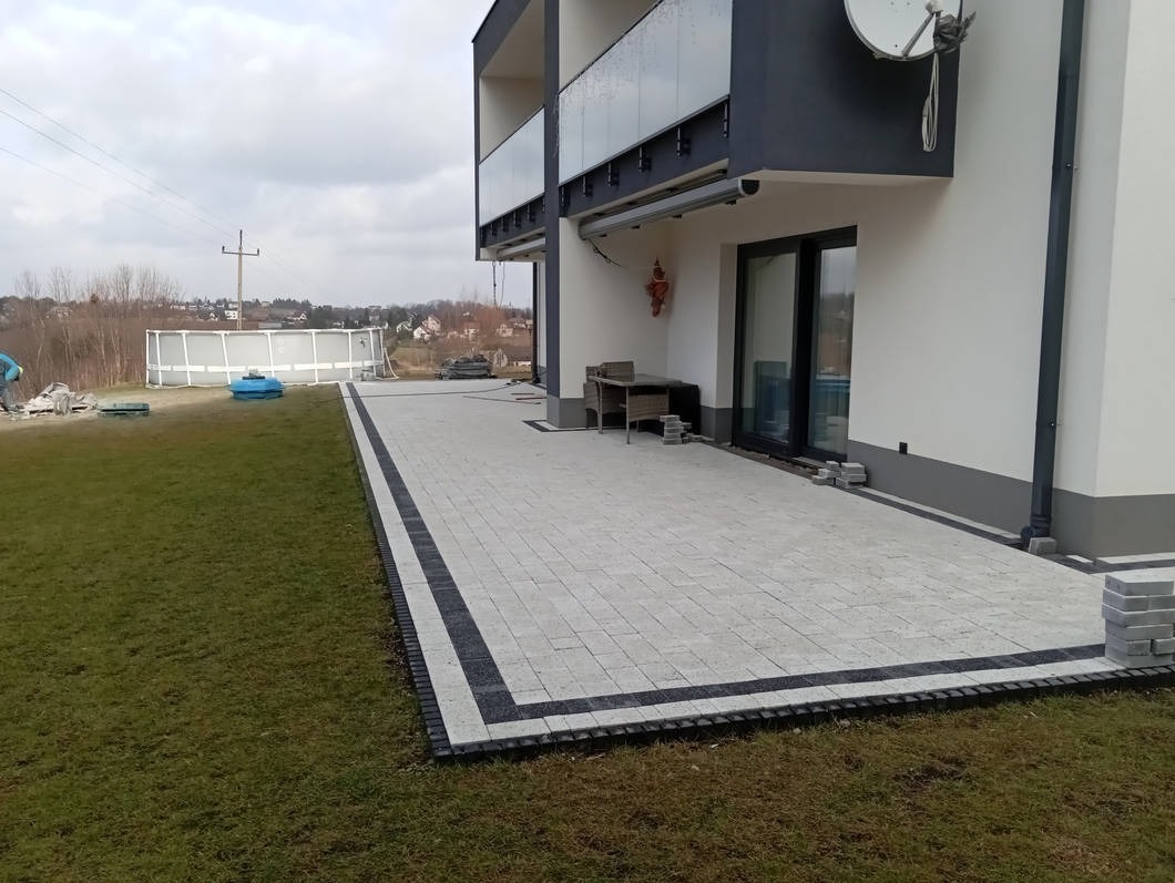 Nowoczesny taras z jasnoszarej kostki z czarną obwódką, przylegający do budynku z balkonami i dużymi oknami tarasowymi. Na trawniku w tle widoczny basen ogrodowy.
