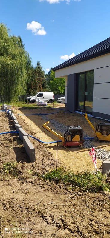Przygotowanie podłoża pod nawierzchnię z kostki brukowej: ubijarki wibrujące na podsypce z kruszywa, obrzeża ustawione wzdłuż wykopu, widoczny budynek z nowoczesną elewacją i zaparkowany samochód...