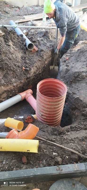 Pracownik w zielonej czapce kopie dół pod rury kanalizacyjne za pomocą szpadla. W wykopie widoczna czerwona rura karbowana, obok różowe i szare rury oraz żółte odcinki rur karbowanych.