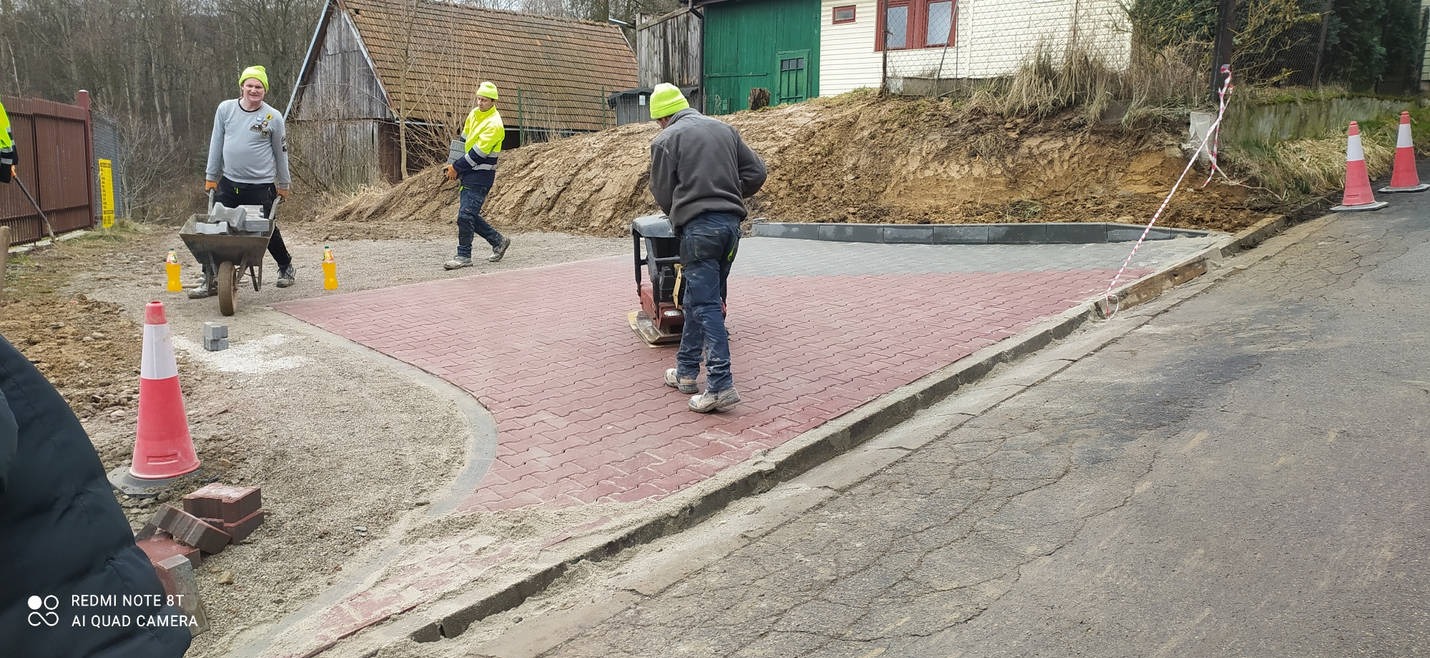Pracownicy w neonowych czapkach układają czerwoną kostkę brukową, używając zagęszczarki; jeden z nich wiezie kostkę na taczce, w tle budynki i usypana ziemia.