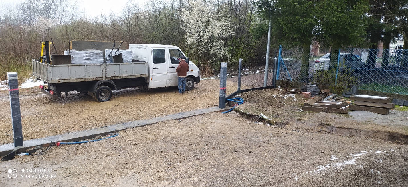 Pracownik przy białym samochodzie dostawczym z kostką betonową, obok przygotowany teren pod układanie kostki z widocznymi słupkami oświetleniowymi i kablem.