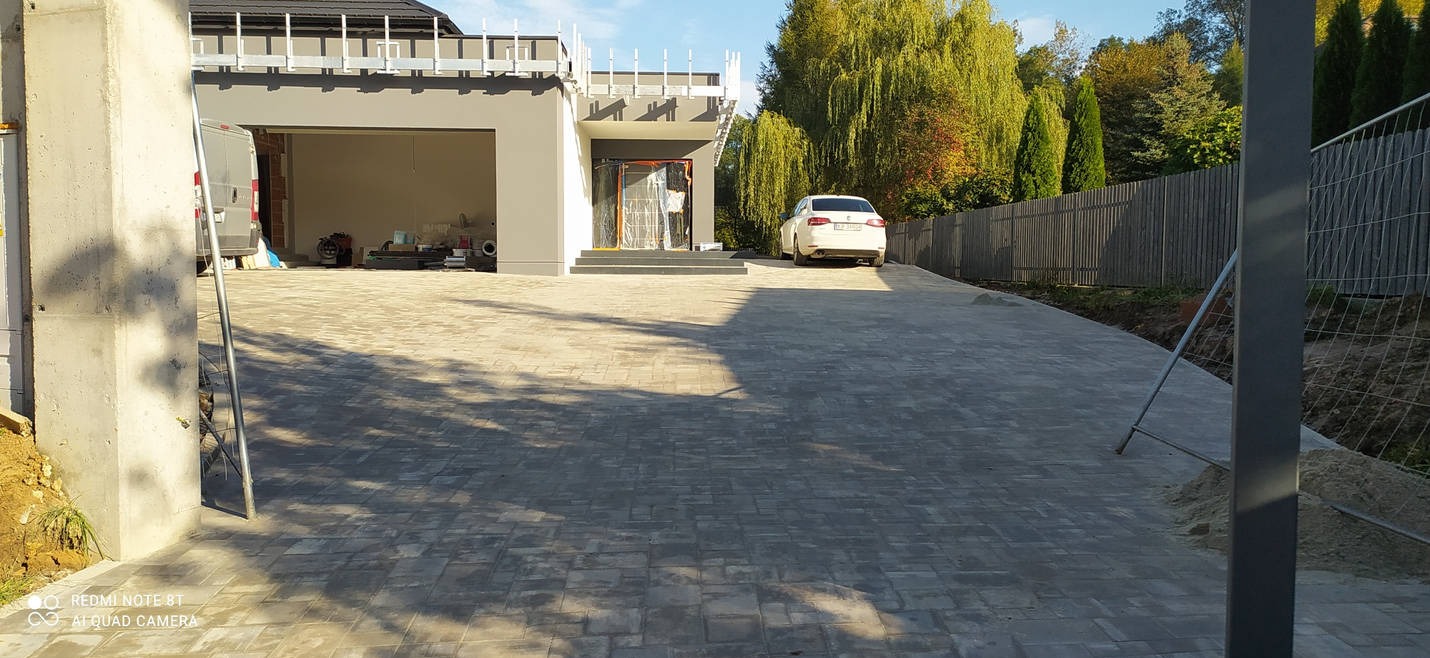 Nowa, szara nawierzchnia z kostki brukowej prowadząca do domu z garażem, widoczny samochód osobowy i ogrodzenie z drewna. Część domu jest jeszcze w folii ochronnej.