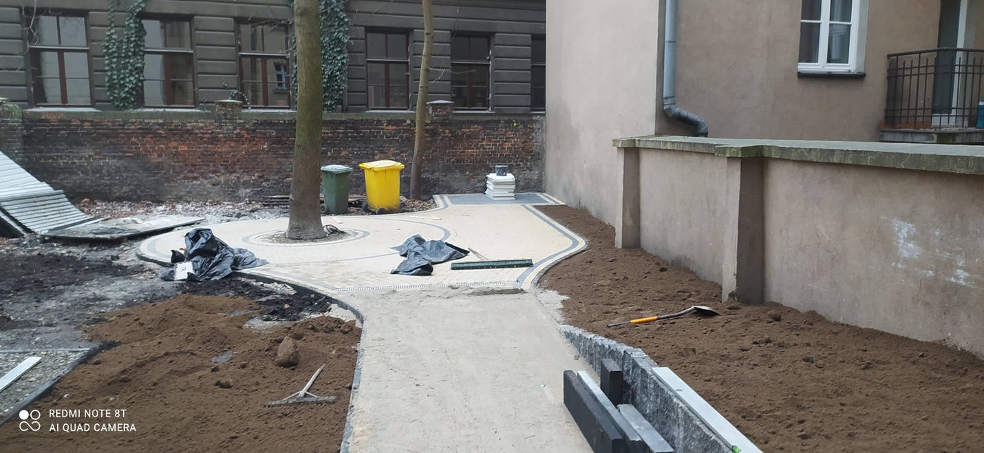 Fragment podwórza w trakcie prac brukarskich: ułożona jasna ścieżka z ciemną obwódką wokół drzewa i wzdłuż muru, obok rozkopana ziemia, widoczne narzędzia i składowane elementy obrzeży.