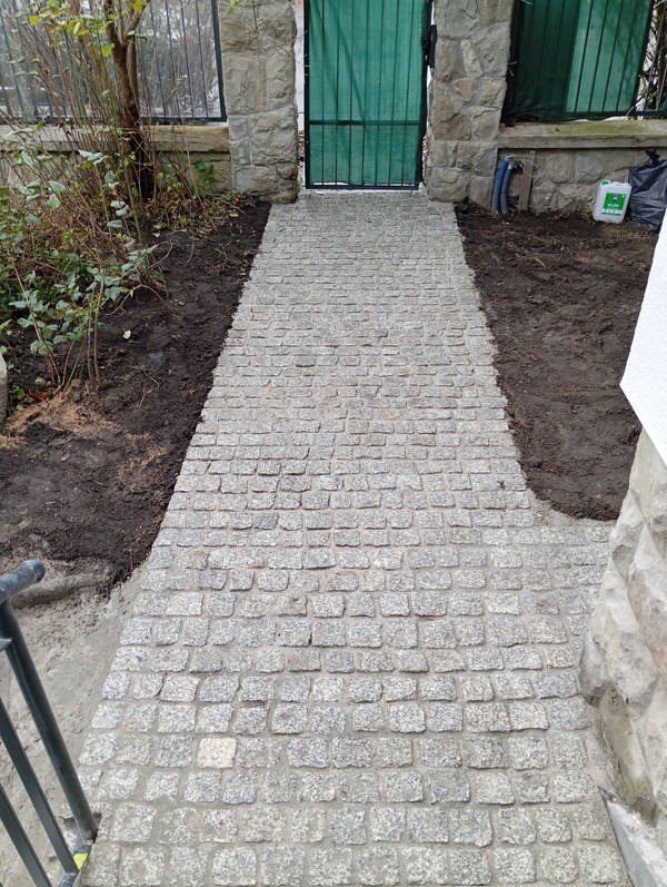 Ścieżka z szarej kostki prowadząca do zielonej bramy, obramowana ziemią i kamiennymi słupkami, widok z góry.