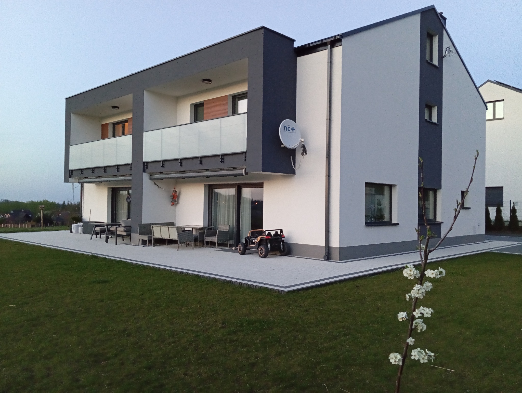 Nowoczesny dwupiętrowy dom z szaro-białą elewacją i tarasem wyłożonym jasną kostką brukową, z meblami ogrodowymi i dziecięcym pojazdem elektrycznym, widziany z perspektywy trawnika z młodym...