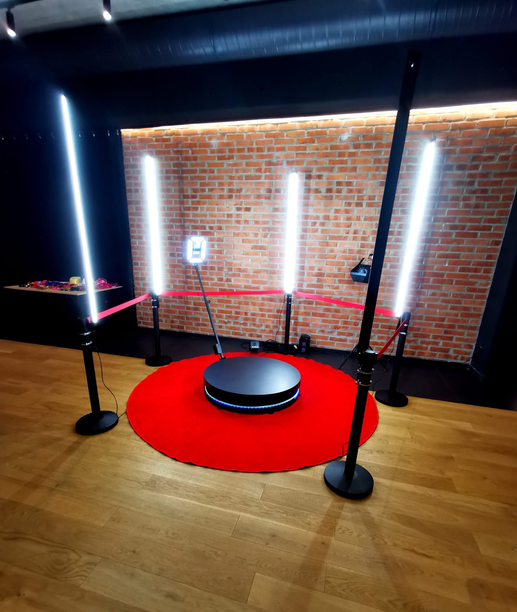 Obrotowa platforma do nagrywania wideo 360 stopni na czerwonym dywanie, otoczona czterema lampami LED na statywach w ciemnym pomieszczeniu z ceglaną ścianą w tle.