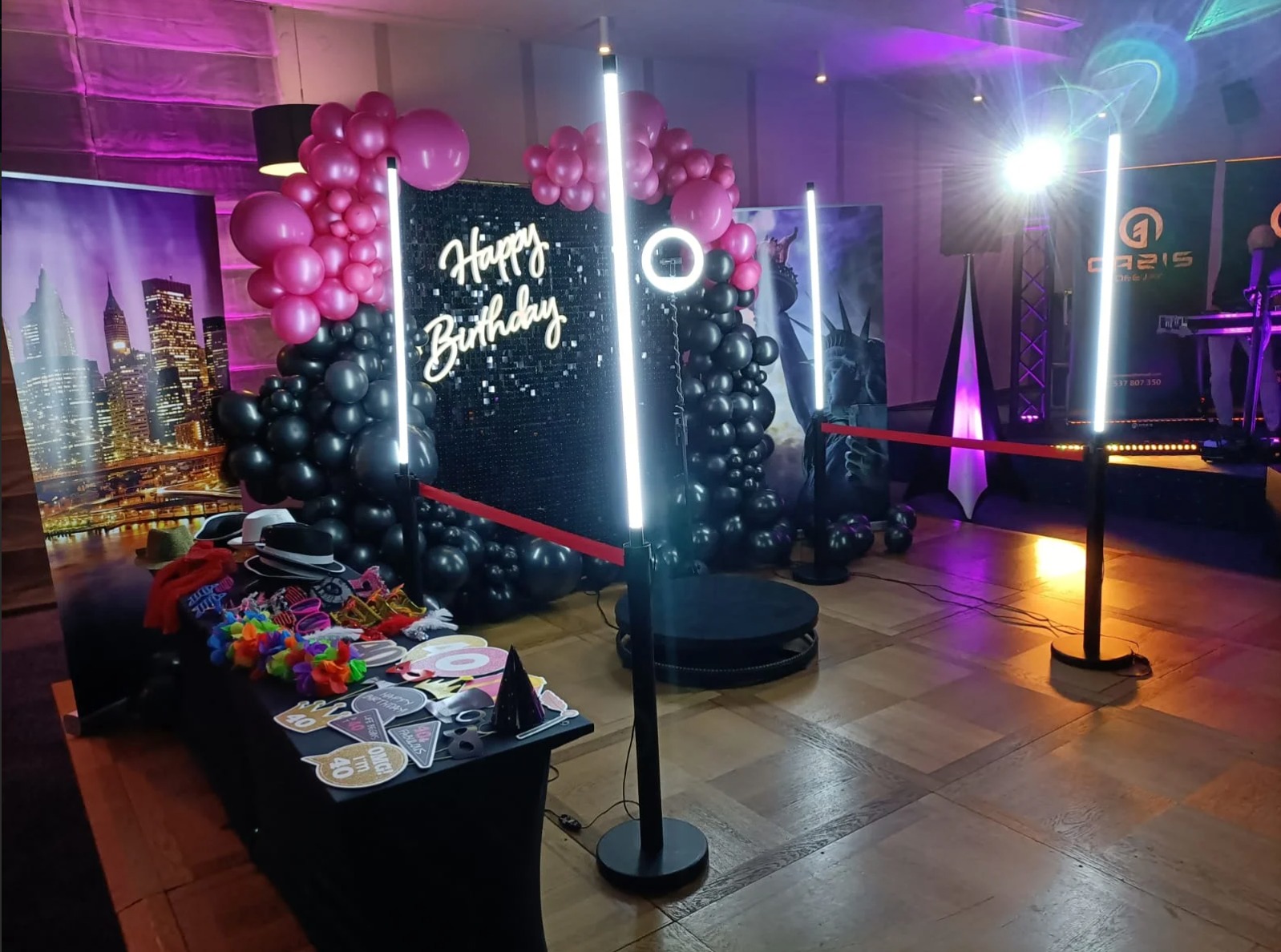 Udekorowana przestrzeń z rekwizytami, balonami różowymi i czarnymi, oraz podświetlanym napisem 'Happy Birthday', przygotowana do robienia zdjęć podczas imprezy.