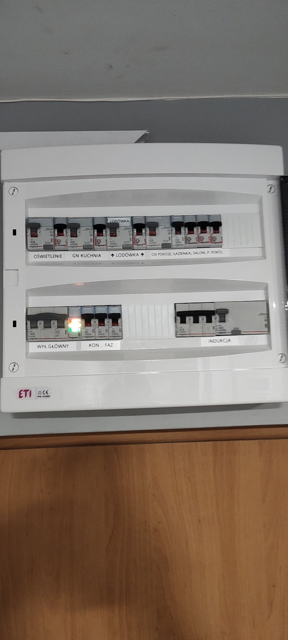 Biała skrzynka rozdzielcza z bezpiecznikami, opisana etykietami: oświetlenie, kuchnia, lodówka, pokoje, indukcja. Jeden bezpiecznik włączony, reszta wyłączona.