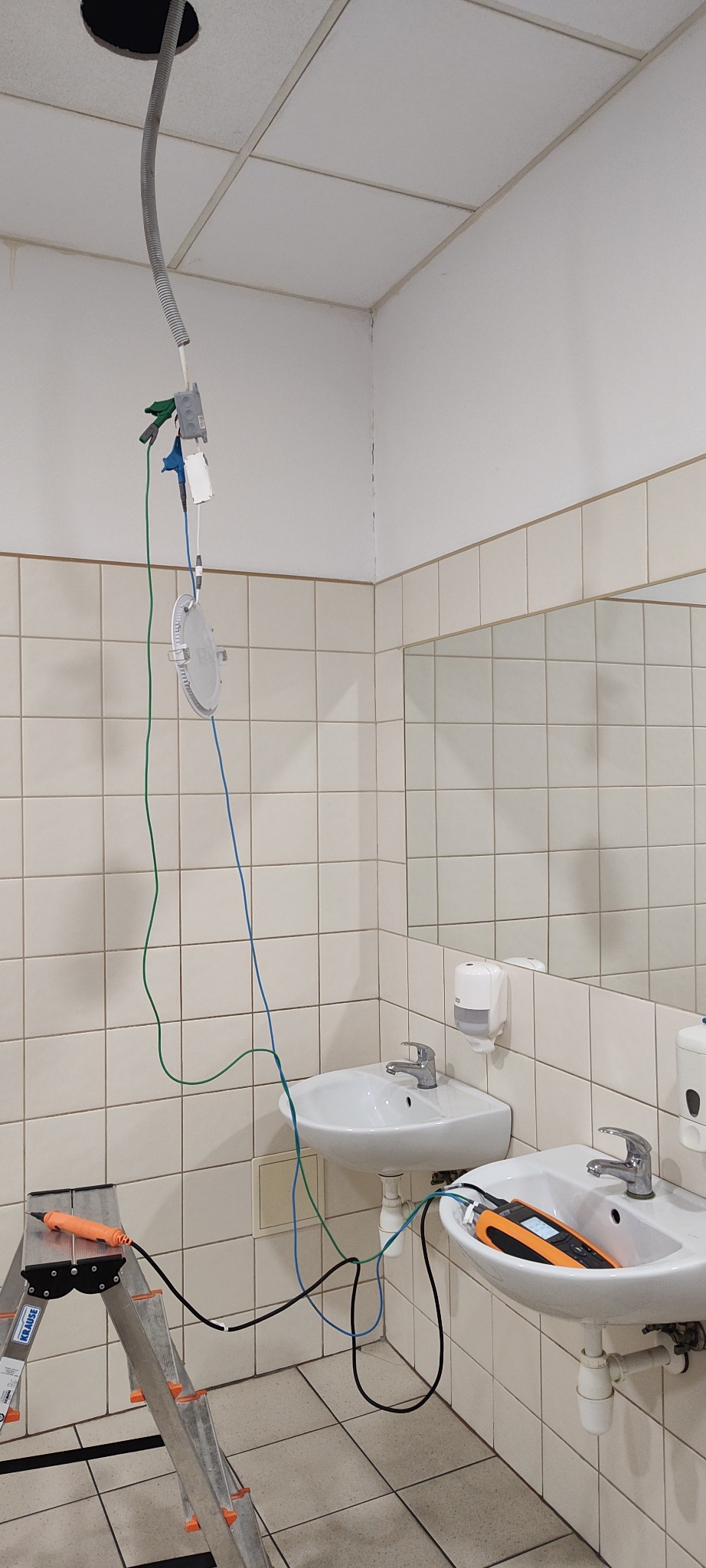 Pomiary elektryczne w łazience: przewody z miernika podłączone do oprawy oświetleniowej zwisającej z sufitu, drabina z narzędziem obok umywalki.