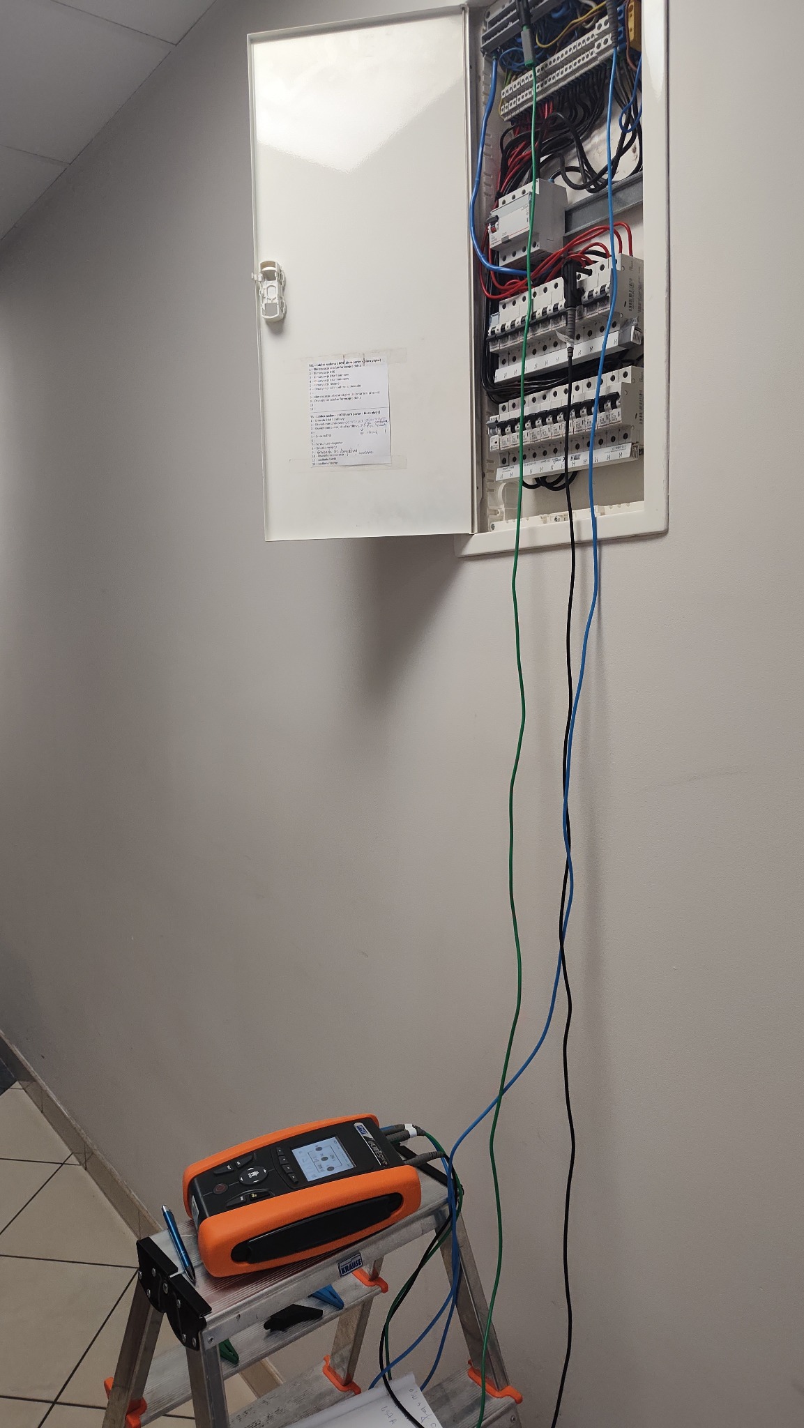 Otwarta skrzynka z bezpiecznikami i przewodami. Na drabinie miernik elektryczny w trakcie pracy. Widoczne kable i schemat. Pomiary instalacji w budynku.