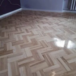 Świeżo położony, lakierowany parkiet ułożony we wzór jodełki francuskiej w pomieszczeniu z szarą i fioletową ścianą, widoczne ślady lakieru na powierzchni.