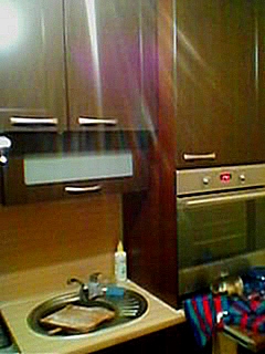 Ciemnobrązowe szafki kuchenne z metalowymi uchwytami nad zlewem z ociekaczem. Widoczny piekarnik w zabudowie i butelka płynu do naczyń.