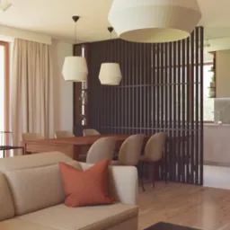 Wnętrze domu jednorodzinnego z beżową sofą, drewnianym stołem i krzesłami, oddzielone ażurową ścianką od kuchni w jasnych barwach.