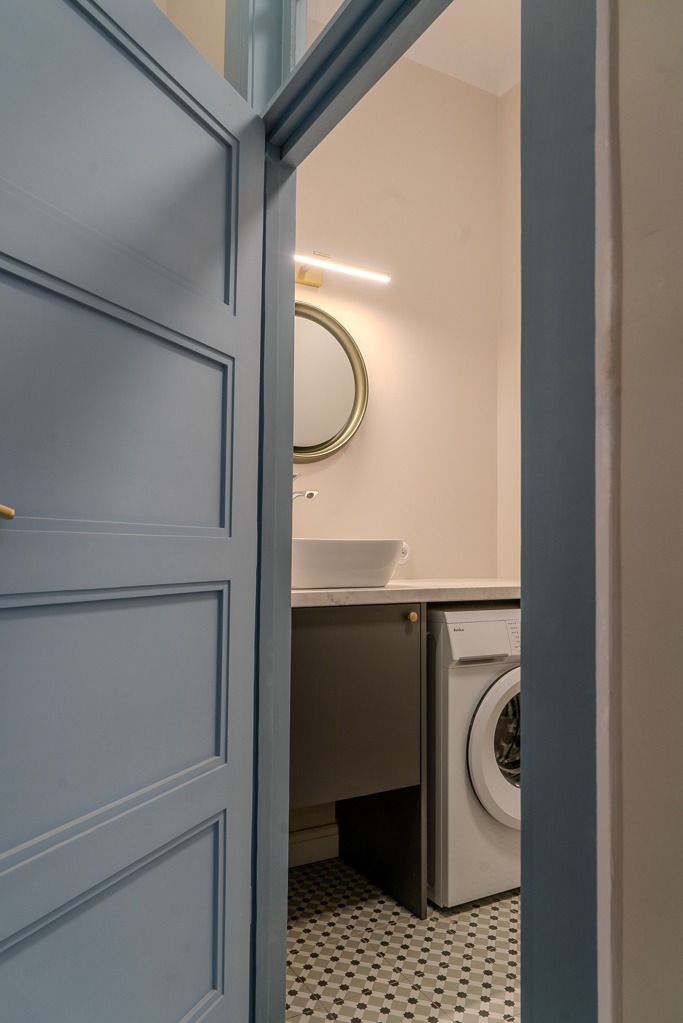 Widok przez uchylone, błękitne drzwi na jasną łazienkę z okrągłym lustrem, umywalką nablatową, szafką pod umywalką i pralką.