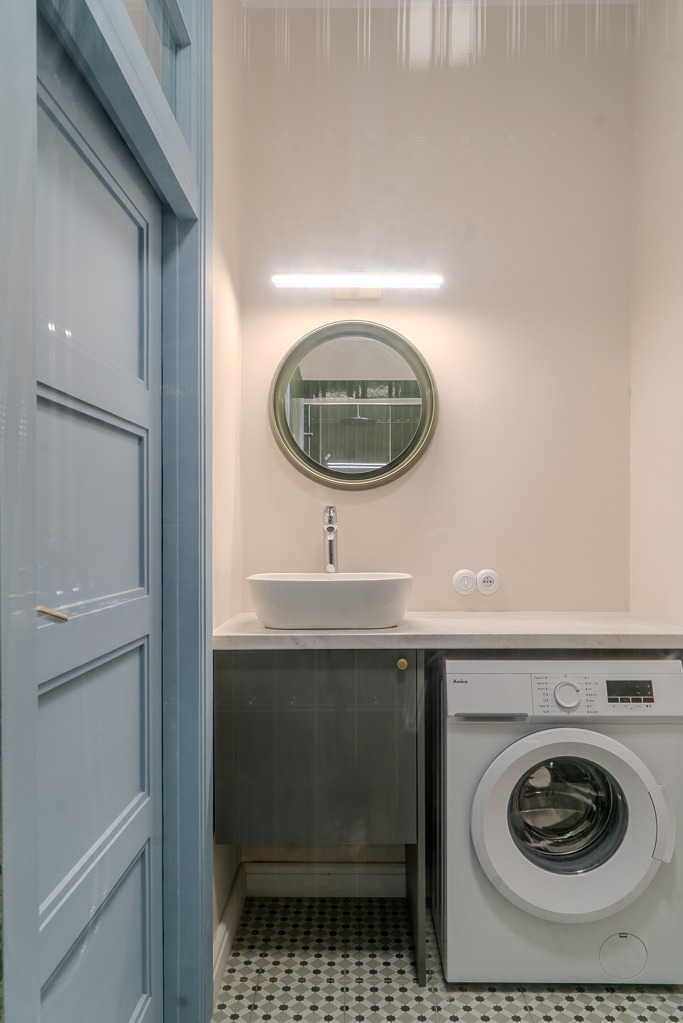 Mała łazienka z umywalką nablatową, okrągłym lustrem w złotej ramie, pralką i szafką pod umywalką w kolorze grafitowym, wykończona mozaiką podłogową.
