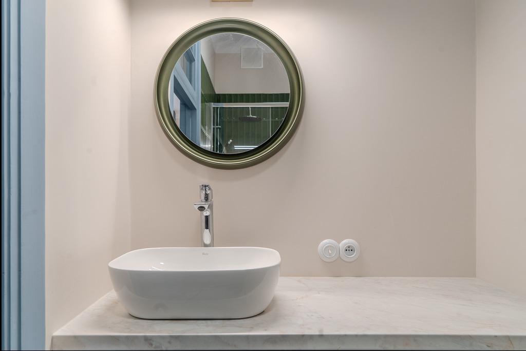 Biała, owalna umywalka na marmurowym blacie, chromowana bateria, okrągłe lustro w zielonej ramie odbijające prysznic z zielonymi płytkami, kremowe ściany i retro gniazdka elektryczne.