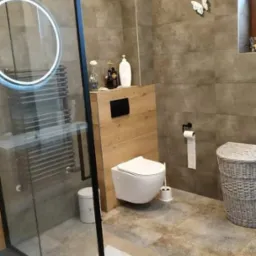 Nowoczesna łazienka z kabiną prysznicową typu walk-in, podwieszaną toaletą i szarą płytką imitującą beton, uzupełniona dekoracyjnymi motylami na ścianie i wiklinowym koszem.