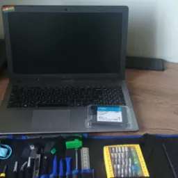 Otwarty laptop marki ASUS z naklejką tęczową w rogu ekranu, dysk SSD Crucial BX500 2.5 na klawiaturze oraz zestaw narzędzi do naprawy elektroniki w rozłożonym etui na drewnianym biurku.
