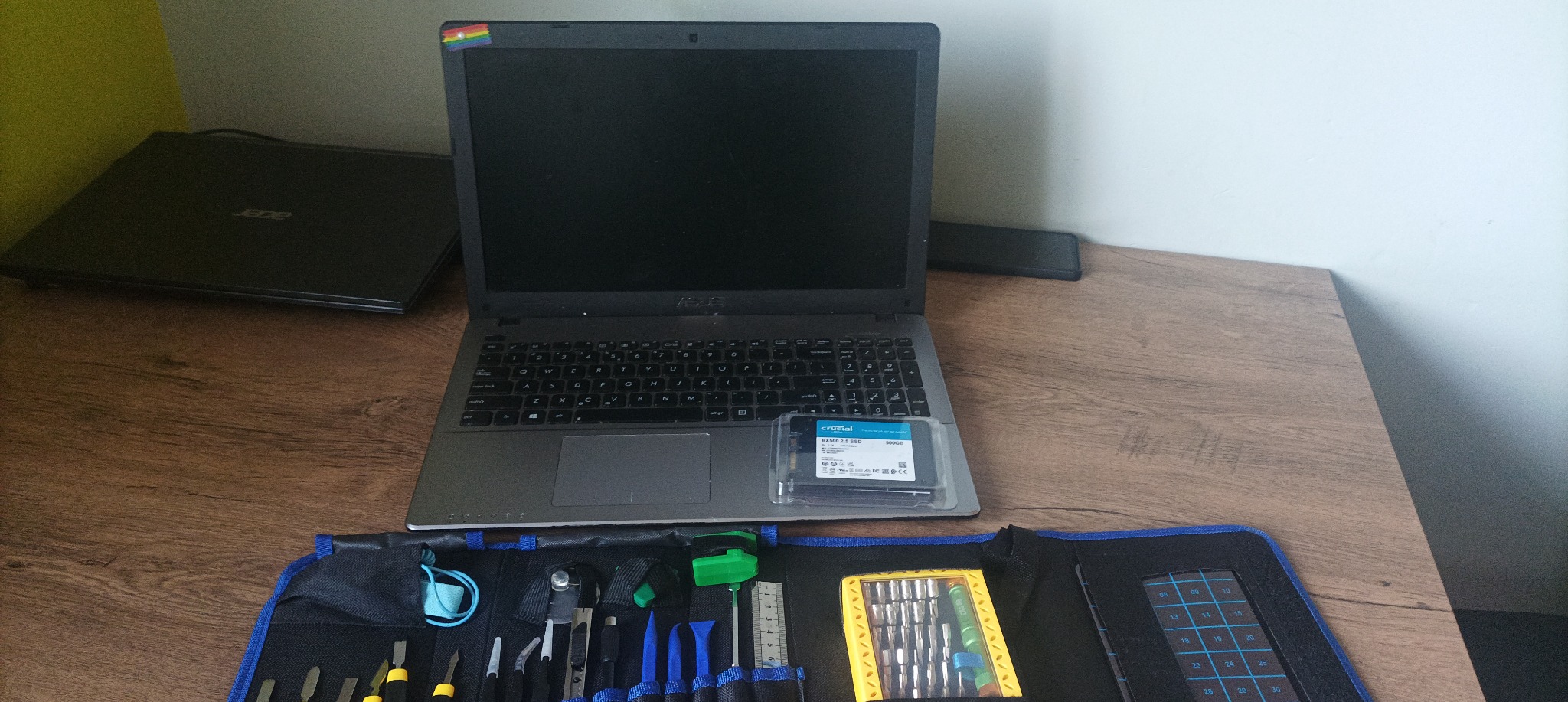 Otwarty laptop marki ASUS z naklejką tęczową w rogu ekranu, dysk SSD Crucial BX500 2.5 na klawiaturze oraz zestaw narzędzi do naprawy elektroniki w rozłożonym etui na drewnianym biurku.