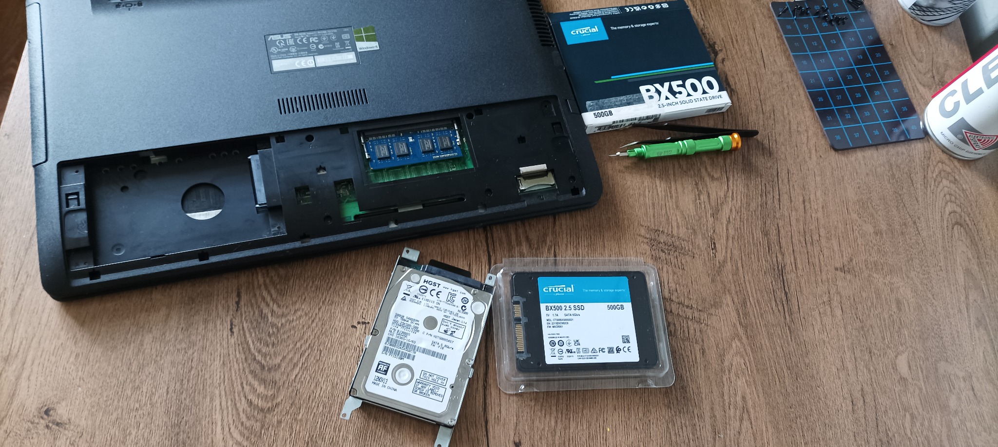 Otwarty laptop z widoczną pamięcią RAM, obok dysk HDD, nowy dysk SSD Crucial BX500 w opakowaniu i luzem, śrubokręt precyzyjny, pudełko po dysku SSD i organizer na śrubki.
