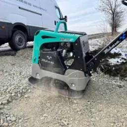 Turkusowo-szara zagęszczarka wibroprasa Mikasa MVH-209 na drodze z kruszywa, w tle biały samochód dostawczy i pole ze śniegiem.