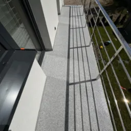 Szara, drobnoziarnista posadzka balkonu z metalową balustradą, widok z góry, w tle trawa i elementy ogrodu, w rogu widoczne narzędzia.