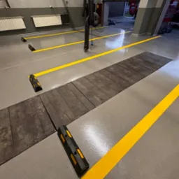 Szara posadzka w warsztacie z żółtymi liniami wyznaczającymi stanowiska, drewnianą platformą i odbojnikami przy stanowiskach.