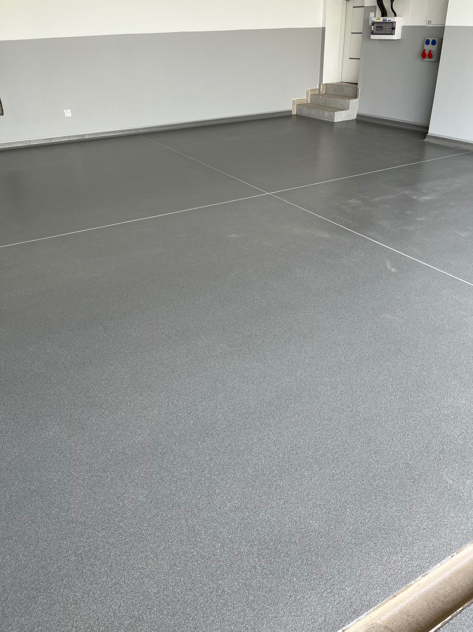 Gładka, szara posadzka z delikatnym połyskiem i widocznymi liniami podziału, w garażu lub pomieszczeniu gospodarczym z szarymi ścianami i białym sufitem.